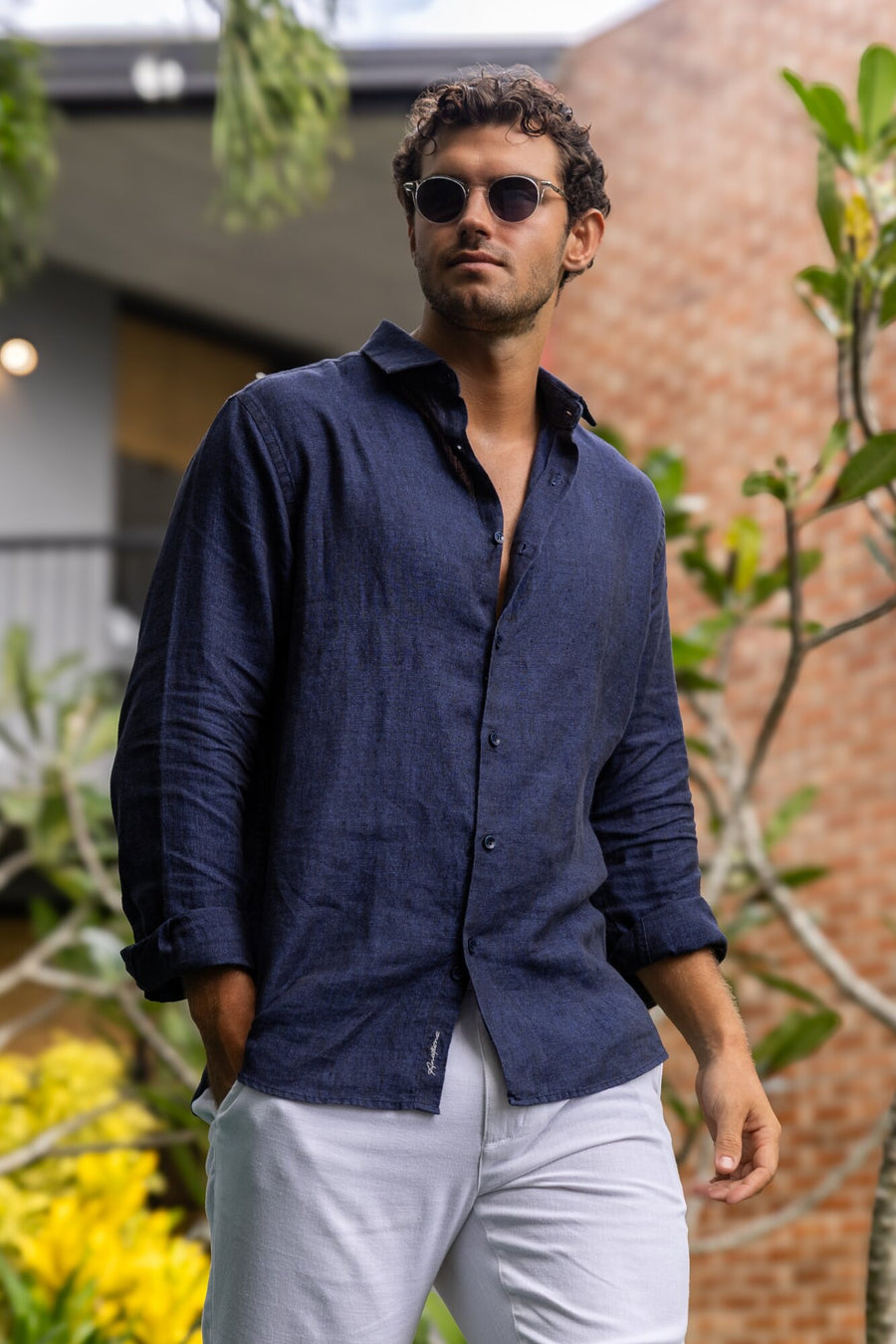 Man in a navy linen shirt and white trousers in a tropical garden